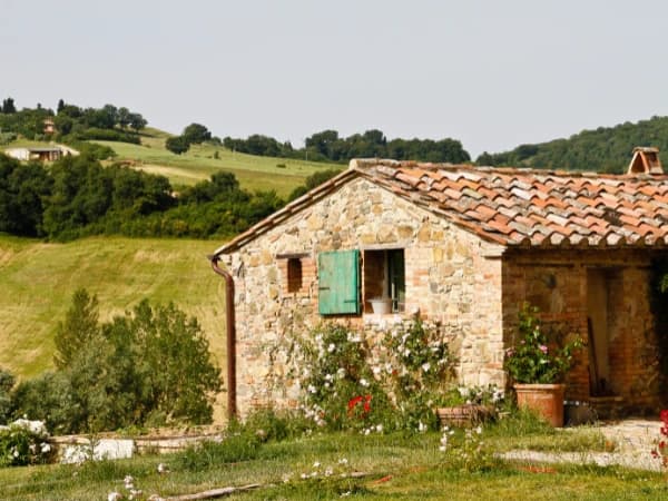 Casa de piedra rústica con tejado de teja y contraventanas verdes, rodeada de vegetación y flores, en un entorno de colinas y campos verdes.